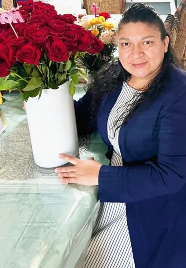 Angela sits at a table holding a big vase of red roses. She has medium skin tone, long dark hair and is wearing a blue blazer over a dress.