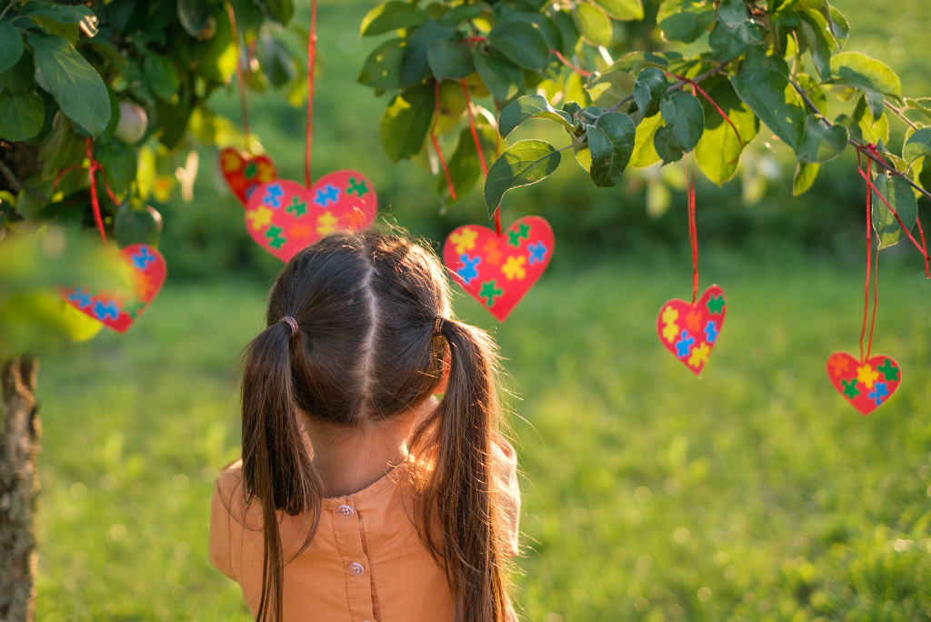 Young child with pigtails faces away from camera and toward a string of red hearts hanging in a tree. Hearts have puzzle pieces on them.