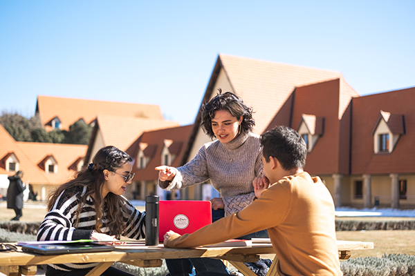 Three students at a picnic bench outside, one with a laptop open