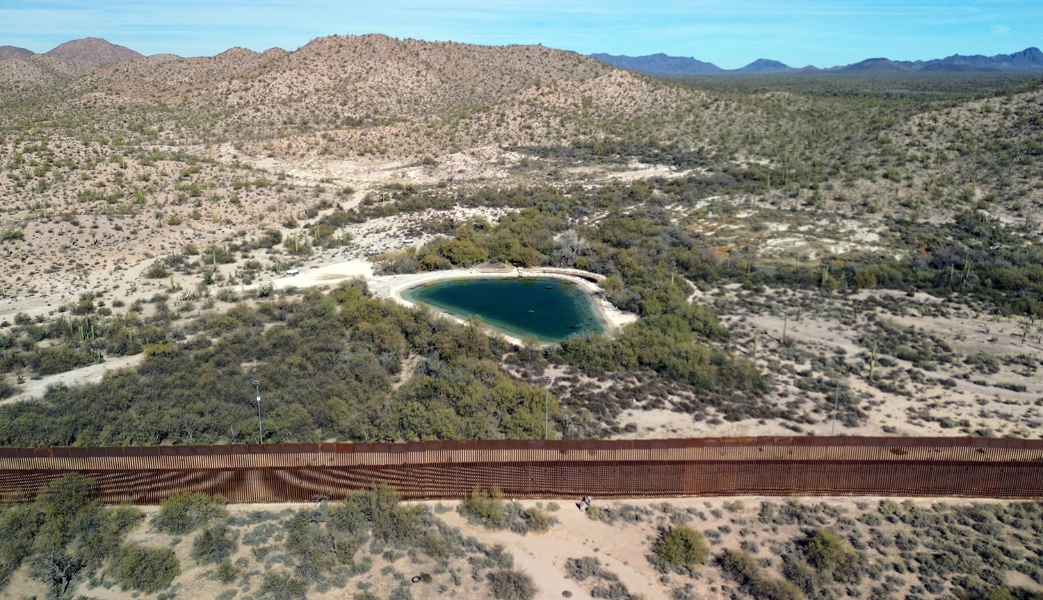 Imagen aérea que muestra un terreno árido, con arbustos verdes y, en el medio, una masa de agua, conocida como el manantial de Quitobaquito. El terreno es atravesado de lado a lado por un muro, impidiendo el acceso al manantial desde el lado sur.