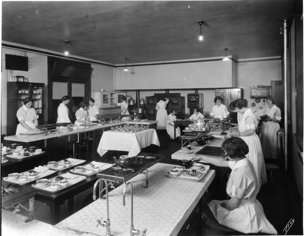 A black and white photo featuring around 10 young women scattered throughout a room that has long white tables with various meal place settings on them, and cupboards along the walls.