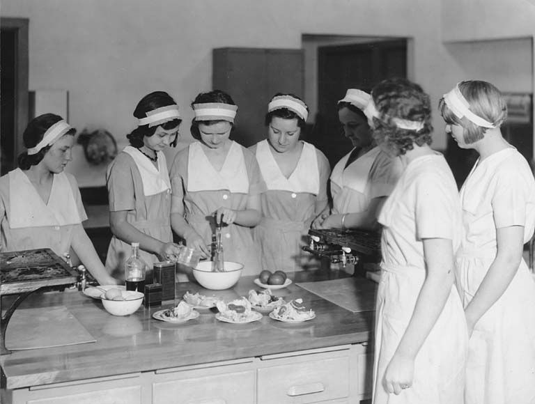 A black a white photo featuring 7 young women with pale faces and dark hair held back with white headbands, all wearing matching dresses with white pinafores. They're standing around a kitchen island watching two of the girls whisking ingredients in a mixing bowl.