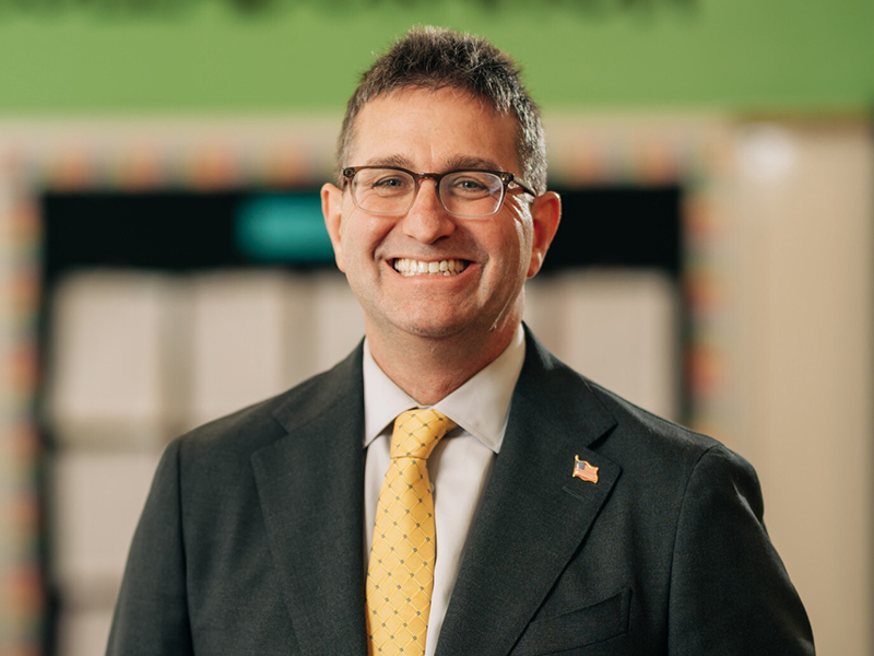 Governor Matt Meyer in a dark suit, white shirt, and yellow tie, wearing glasses and smiling. He has short dark hair with some gray, and an American flag lapel pin on his suit. The background features a blurred classroom setting with a green wall and bulletin boards.