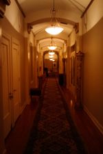 Looking along a hallway with a long geometric carpet over a wood floor, arches and columns spaced every so often, a few decorative columns against the walls, all dimly lit by yellowish, dish-shaped lamps hanging from the ceiling.