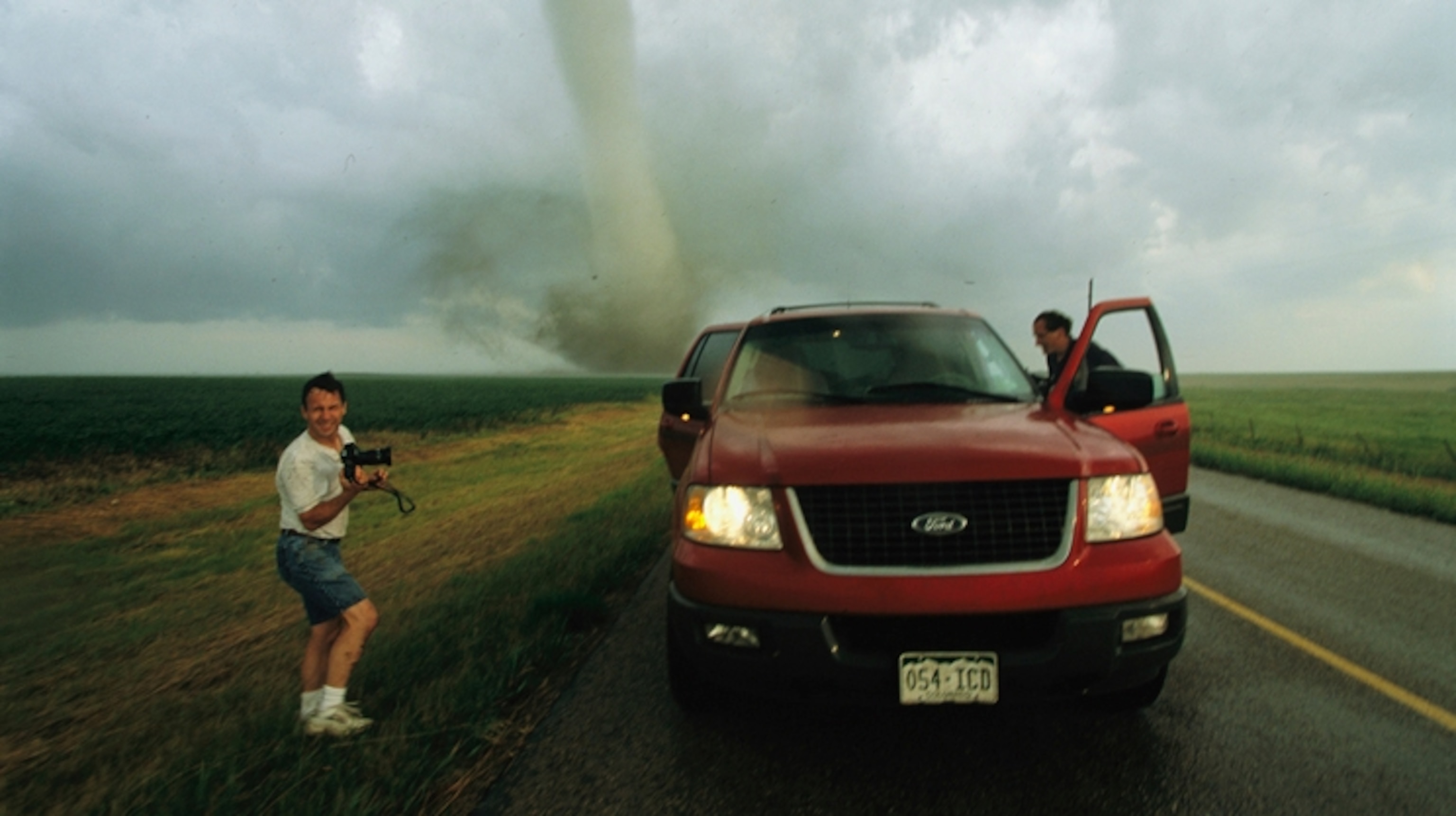 Two men near a red truck with a tornado in the background