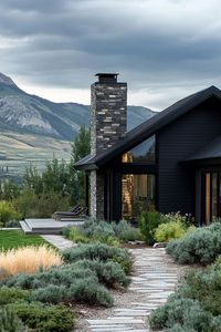 Modern cottage style mountain house with dark stone foundation and chimney black siding large panoramic windows landscape with shrubs and stone. Check out all of these inspiring modern dark mountain houses that blend sleek design with nature’s rugged beauty for an exquisite home.