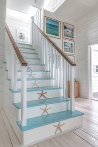 The staircase reflects coastal farmhouse decor with its wooden steps painted in light blue and white. Starfish and seashell motifs on the risers add a playful, maritime touch. A rope handrail further emphasizes the nautical theme, while framed family photos on the wall create a personal gallery. The space is functional yet decorative, serving as both a transition area and a focal point of the home. Click to discover more coastal farmhouse staircase designs!