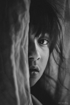 a black and white photo of a young woman peeking out from behind a sheer curtain