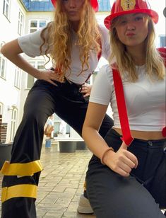 two women wearing fireman hats and suspenders posing for the camera with their hands on their hipss