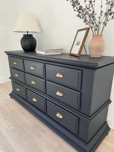 a black dresser with gold handles and drawers in a white walled room next to a lamp