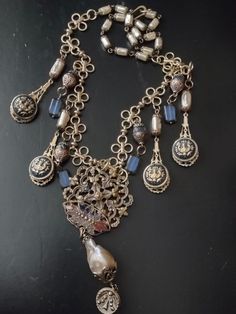 An assemblage featuring a French unmarked antique buckle half with traces of old enamel. It suspends a large baroque freshwater pearl and an antique Victorian cut steel button. The baroque pearl is missing a patch of nacre on the back, but is still luminous and good quality. Accompanying pendants are vintage style blue enameled brass buttons set on brass filigree with faceted iolite, blue Czech glass rectangles and brass capped Czech glass pearls. Finishes with Czech glass pearl chain and hook. One Of A Kind Antique Jewelry, Ornate Metal Jewelry Vintage Collection, Ornate Metal Jewelry For Vintage Collection, Antique Bronze Decorative Jewelry, Antique Gold Decorative Jewelry, Ornate Handmade Jewelry For Vintage Occasions, Handmade Ornate Vintage Jewelry, Victorian Vintage Charm Metal Jewelry, Vintage Baroque Jewelry For Formal Occasions