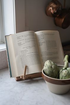 an open book and some artichokes in a bowl on a marble counter