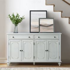 a white sideboard with two pictures on it next to a stair case and potted plant