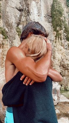 a man and woman hugging each other in front of a cliff face by the water