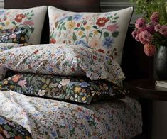 a bed topped with lots of pillows next to a vase filled with pink and purple flowers