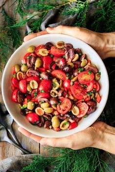 two hands holding a white bowl filled with olives and tomatoes