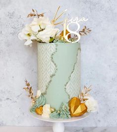 a green cake with gold and white flowers on top is sitting on a pedestal in front of a marble wall