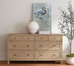 a white vase sitting on top of a wooden dresser next to a painting and a potted plant