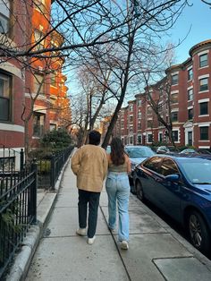 two people walking down the sidewalk in front of parked cars and apartment buildings on a sunny day