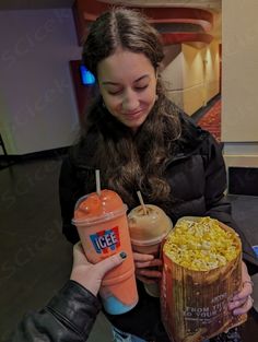 a woman holding two drinks and a box of popcorn