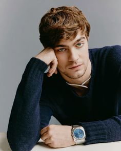 a young man wearing a blue sweater leaning on a table with his hand on his head