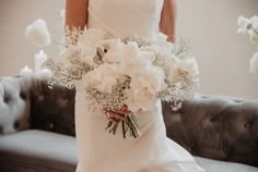 a bride holding a bouquet of white flowers in her wedding dress and standing next to a couch