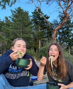 two girls eating food out of green bowls on the ground in front of some trees