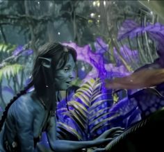 a woman with blue paint on her face and body in front of purple plants, surrounded by leaves