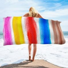 a woman standing on the beach holding a rainbow colored towel over her head and looking out at the ocean