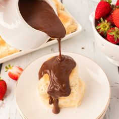 chocolate sauce being poured onto a biscuit on a white plate with strawberries in the background