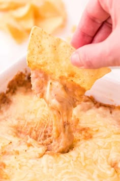 a hand holding a tortilla chip over a casserole dish