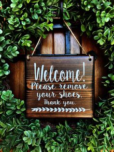 a wooden sign hanging from the side of a green bush with leaves around it that says, welcome please remove your shoes thank you