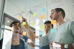 a group of people standing around a glass wall with sticky notes on it and one man writing