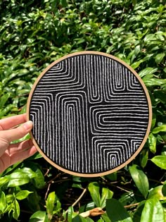 a hand holding up a black and white pattern on a wooden frame in front of some bushes