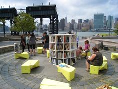 people are sitting and standing around bookshelves near the water in front of a cityscape