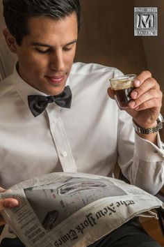 a man in a white shirt and bow tie reading a newspaper while holding a glass of beer