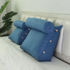 three blue pillows on a white bed with a green plant in the corner behind them