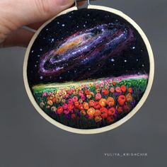 a hand holding up a small embroidery art piece with flowers and the milky in the background