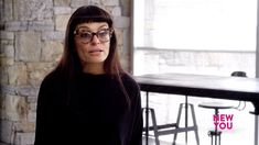 a woman wearing glasses standing in front of a table with chairs and stools behind her