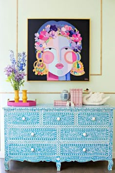 a painting on the wall above a blue dresser with flowers in front of it and vases