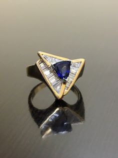 DeKara Designs Clearance Metal- 14K Yellow Gold, .583. 15.6 Grams. Stones- 1 Trillion Cut Ceylon Blue Sapphire 0.85 Carats, 21 Tapered Baguette Diamonds G-H Color VS2 Clarity 0.90 Carats. Size- Ring is a 6. FREE SIZING Art Deco Inspired Handmade 14K Yellow Gold Ceylon Blue Sapphire Diamond Ring. This ring is inspired by the Art Deco era with unique Trillion Cut Ceylon Blue Sapphire that is set in between three V prongs. There are 21 professionally channel set tapered baguette diamonds surroundin Blue Tanzanite Ring With Trillion Cut, Gia Certified Blue Art Deco Jewelry, Blue Art Deco Jewelry Gia Certified, Trillion Cut Sapphire Ring In 14k Gold, 14k Gold Trillion Cut Sapphire Ring, Fine Jewelry Yellow Gold Trillion Cut Sapphire Ring, Trillion Cut Sapphire Rings For Formal Occasions, Blue Art Deco Jewelry Stamped 14k, Elegant Blue Trillion Cut Diamond Ring