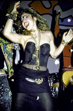 a woman in a black dress dancing with her hands up and arms outstretched, wearing gold jewelry