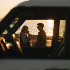 a man and woman sitting on the beach in front of a car looking at each other