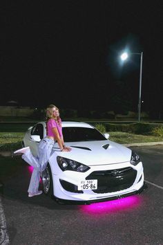 a woman leaning on the hood of a white car in a parking lot at night