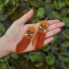 a pair of orange beaded earrings on a person's hand with green plants in the background