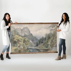 two women standing next to each other holding up a painting with mountains in the background