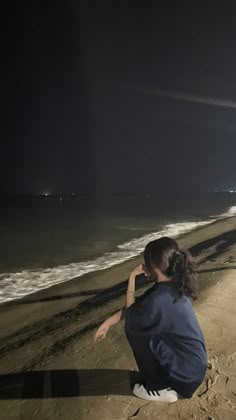 a woman sitting on top of a sandy beach next to the ocean under a dark sky