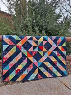 a colorful quilt sitting on top of a cement slab next to trees and bushes in the background