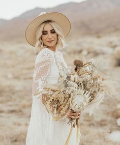 a woman in a white dress and hat holding a bunch of dried flowers with mountains in the background