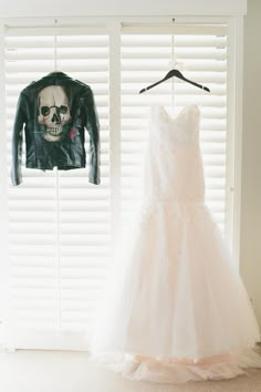 a wedding dress hanging in front of a leather jacket on a hanger next to a window